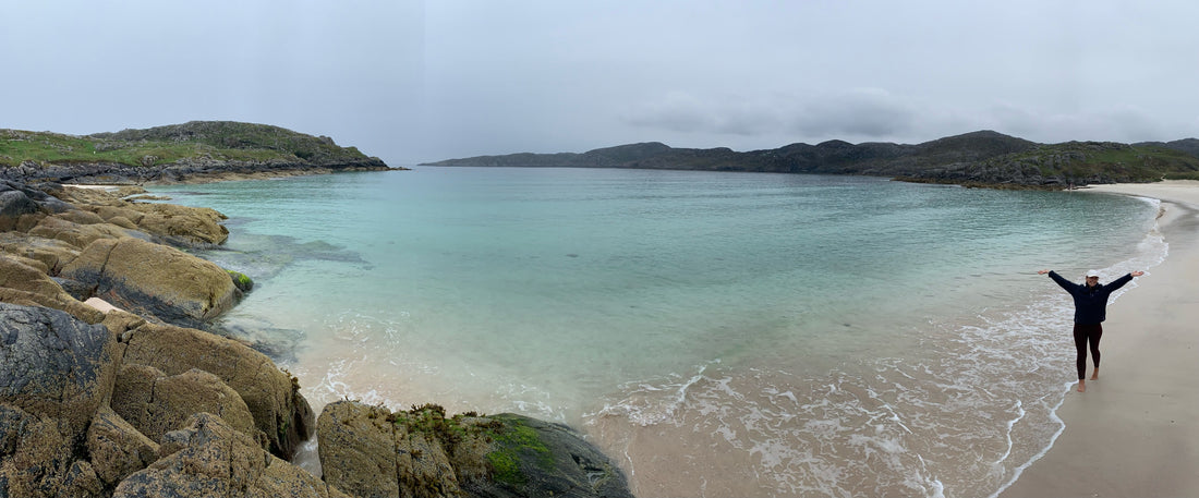 Woman posing with arms in the air next to a white sand, crystal clear Scottish beach